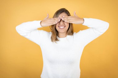 İzole edilmiş sarı arka planda genç beyaz kadın, neşeli ve komik gülümseyen ellerle gözlerini kapatıyor. Kör kavram.