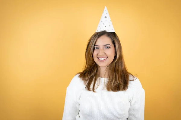 Young caucasian wearing a birthday hat woman over isolated yellow background with a happy face standing and smiling with a confident smile showing teeth