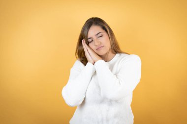 Kafkasyalı genç bir kadın izole edilmiş sarı arka planda uyuyor. Yorgun düşmüş ve elleriyle poz veriyor, gözleri kapalı gülümsüyor..