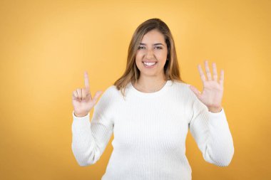 İzole edilmiş sarı arka planda genç beyaz kadın yedi numaralı parmaklarıyla gülümseyerek kendinden emin ve mutlu görünüyor.