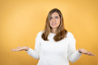 İzole edilmiş sarı arka plan üzerinde beyaz bir kadın. Kolları ve elleri havada, kafası karışmış bir ifade.