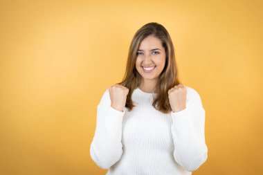 Genç beyaz kadın izole edilmiş sarı arka planda çok mutlu ve heyecanlı kollarını kaldırarak zafer jesti yapıyor, gülümsüyor ve başarı için bağırıyor..