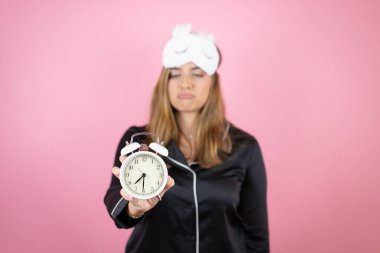 İzole edilmiş pembe arka plan üzerinde uyku maskesi ve pijama giyen genç beyaz kadın uyuyor ve saati gösteriyor.