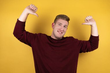 Sarı arka planda sıradan kırmızı bir kazak giyen genç sarışın. Kendine güvenen, yüzünde gülümseme olan, kendini gururlu ve mutlu gösteren bir adam..