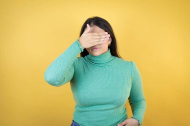 Kafkasyalı genç bir kadın izole edilmiş sarı arka planda ciddi ve elleriyle gözlerini kapatıyor.