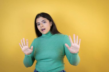 Kafkasyalı genç bir kadın sarı arka planda korku ve korku ifadesinden korkuyor ve dehşete düşüyor. El hareketlerini durdur, şok içinde bağır. Panik konsepti.