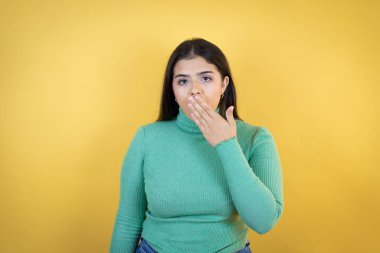 İzole edilmiş sarı arka planda genç beyaz kadın ağzını kapatırken şaşırdı.