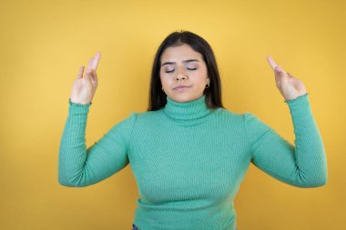 İzole edilmiş sarı arka plan üzerinde genç beyaz kadın rahatla ve gözleri kapalı gülümse parmaklarıyla meditasyon hareketi yapıyor.