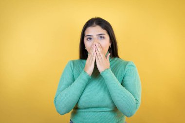 Kafkasyalı genç bir kadın, izole edilmiş sarı arka planda, elleri ağzında ve şaşırmış bir şekilde.