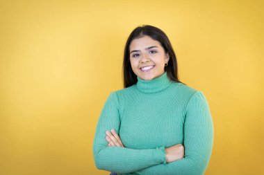 Genç beyaz kadın, izole edilmiş sarı arka planda, mutlu bir yüzü var, ayakta duruyor ve gülümsüyor, kollarını kavuşturmuş dişlerini gösteriyor.