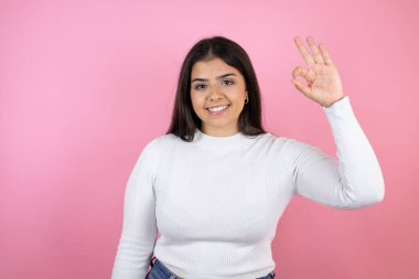 Soyutlanmış pembe arka planda genç ve güzel bir kadın parmaklarıyla imza atıyor ve gülümsüyor, mükemmel bir sembol.