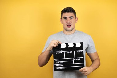 Sarı arka planda sıradan bir tişört giyen yakışıklı bir genç. Elinde klaket tutarken çok mutlu.