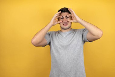 Sarı arka plan üzerine sıradan bir tişört giyen genç yakışıklı adam parmaklarıyla gözlerini açmaya çalışıyor, uykulu ve yorgun bir şekilde sabah yorgunluğu çekiyor.