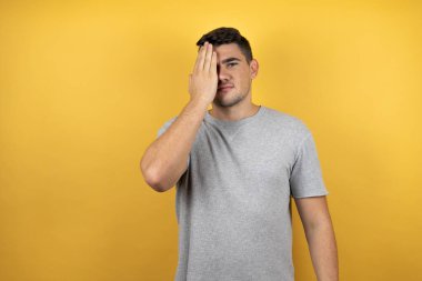Sarı arka plan üzerine sıradan bir tişört giyen genç yakışıklı bir adam bir gözünü eliyle kapatıyor, yüzünde kendine güvenen bir gülümseme ve sürpriz bir duygu var..