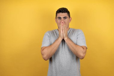 Genç ve yakışıklı bir adam, sarı arka planda, elleri ağzında, günlük bir tişört giyiyor ve şaşırıyor.