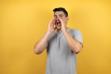 Sarı arka planda rahat bir tişört giyen genç yakışıklı adam elleri ağızda bağırarak ve yüksek sesle bağırıyor.