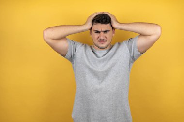 Sarı arka plan üzerine sıradan bir tişört giyen genç yakışıklı adam baş ağrısı ve stresten muzdarip çünkü elleri başının üstünde acı ve migren var.