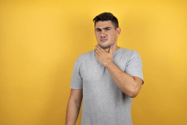 İzole edilmiş sarı arka plan üzerine günlük tişört giyen genç yakışıklı adam ağrılı boyuna dokunuyor, grip için boğaz ağrısı, mantar ve enfeksiyon kapıyor.
