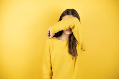 Pretty girl wearing a yellow sweater standing over isolated yellow background covering eyes with arm smiling cheerful and funny. Blind concept.
