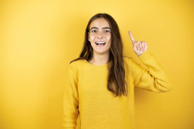 Pretty girl wearing a yellow sweater standing over isolated yellow background having an idea
