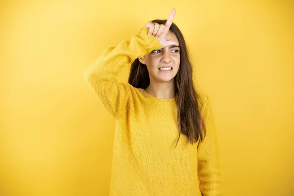 Pretty girl wearing a yellow sweater standing over isolated yellow background making fun of people with fingers on forehead doing loser gesture mocking and insulting.