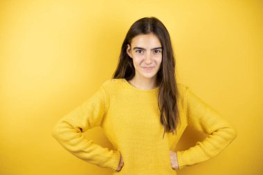 Pretty girl wearing a yellow sweater standing over isolated yellow background skeptic and nervous, disapproving expression on face with arms in waist
