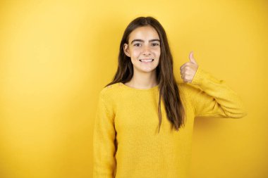 Pretty girl wearing a yellow sweater standing over isolated yellow background smiling confident doing the ok signal with her thumbs