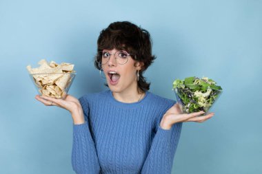 Young beautiful woman holding nachos and healthy salad over isolated blue background afraid and shocked with surprise and amazed expression, fear and excited face.