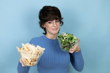 Young beautiful woman holding nachos and healthy salad over isolated blue background smiling looking to the side and staring away thinking.