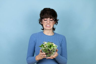 Young beautiful woman holding holding bowl with salad over isolated blue background disgusted expression, displeased and fearful doing disgust face because aversion reaction. Annoying