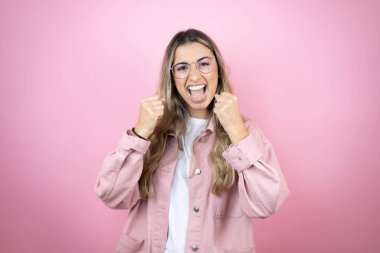 Pembe arka planda duran güzel sarışın kadın çok mutlu ve heyecanlı. Kollarını kaldırmış, gülümseyen ve başarı için çığlık atan kazanan bir jest yapıyor..