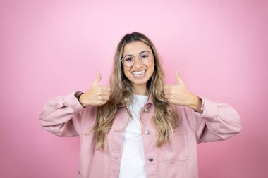 Pembe arka planda duran uzun saçlı güzel sarışın kadın gülümsüyor ve baş parmaklarıyla işaret veriyor.