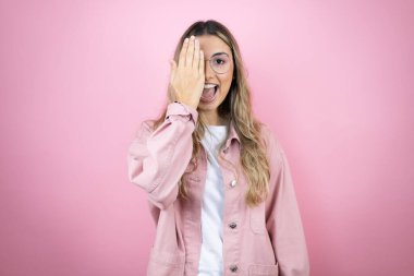 Uzun saçlı güzel bir kadın pembe arka planda duruyor bir gözünü eliyle kapatıyor, yüzünde kendine güvenen bir gülümseme ve sürpriz bir duygu var..