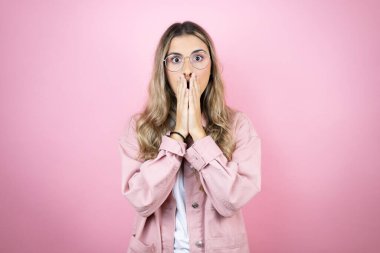 Genç, sarışın, uzun saçlı, pembe arka planda duran, elleri ağzında ve şaşırmış genç bir kadın.