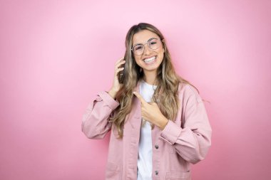Pembe arka planda duran uzun saçlı güzel sarışın kadın gülümsüyor ve telefonu işaret ediyor.