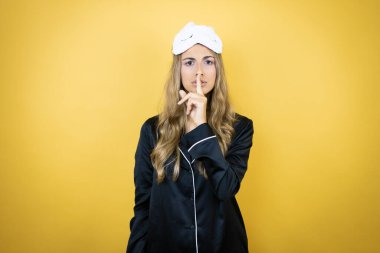 İzole edilmiş sarı arka plan üzerinde uyku maskesi ve pijama giyen genç ve güzel bir kadın sessiz olmak istiyor. Sessizlik ve gizli konsept.