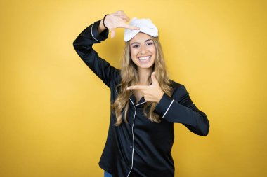 İzole edilmiş sarı arka plan üzerinde uyku maskesi ve pijama giyen genç ve güzel bir kadın gülümsüyor elleri ve parmakları mutlu yüzlü bir çerçeve yapıyor.