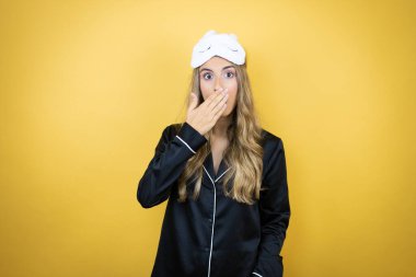 İzole edilmiş sarı arka plan üzerinde uyku maskesi ve pijama giyen güzel bir kadın ağzını kapatırken şaşırdı.