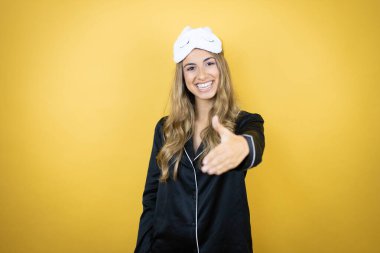 İzole edilmiş sarı arka plan üzerinde uyku maskesi ve pijama giyen genç ve güzel bir kadın dostça bir selamlama ve hoş geldin hediyesi olarak el sıkışma sunuyor.