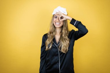 İzole edilmiş sarı arka plan üzerinde uyku maskesi ve pijama giyen genç ve güzel bir kadın gülümseyen yüzle, gözleriyle parmaklarından bakarak şok oluyor.