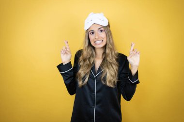 İzole edilmiş sarı arka plan üzerinde uyku maskesi ve pijama giyen genç ve güzel bir kadın umut dolu gülümseyerek ve etrafa bakarak el sallıyor.