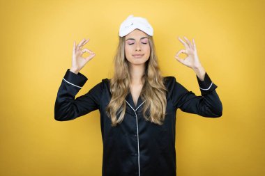 İzole edilmiş sarı arka plan üzerinde uyku maskesi ve pijama giyen genç ve güzel bir kadın rahatla ve gözleri kapalı gülümse parmaklarıyla meditasyon hareketi yapıyor.