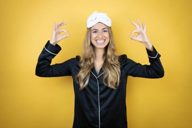 İzole edilmiş sarı arka plan üzerinde uyku maskesi ve pijama giyen genç güzel bir kadın tamam işareti yapıyor parmaklarıyla ve gülümsemesiyle, mükemmel bir sembol.