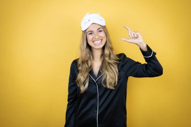 İzole edilmiş sarı arka plan üzerinde uyku maskesi ve pijama giyen genç ve güzel bir kadın gülümsüyor ve el hareketleriyle parmaklarıyla küçük boy işareti yapıyor. Ölçü kavramı.