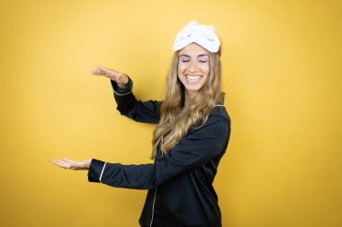 İzole edilmiş sarı arka plan üzerinde uyku maskesi ve pijama giyen genç ve güzel bir kadın. Ellerinde büyük ve büyük boy işareti, ölçü sembolü var. Gülümsüyor