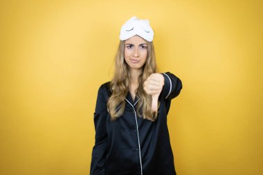 İzole edilmiş sarı arka plan üzerinde uyku maskesi ve pijama giyen genç ve güzel bir kadın. Kızgın bir yüzü var.