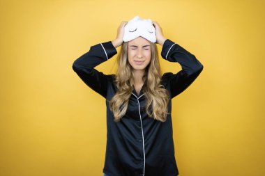 Uyku maskesi ve pijama giyen genç ve güzel bir kadın. Sarı arka plan üzerinde. Baş ağrısı, çaresizlik ve stresten muzdarip.