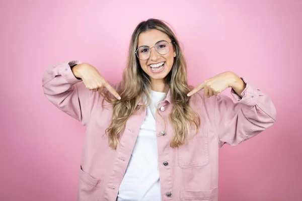 Pembe arka planda duran güzel sarışın kadın. Kendine güvenen, gülümseyen, kendini gururlu ve mutlu parmaklarıyla gösteren..