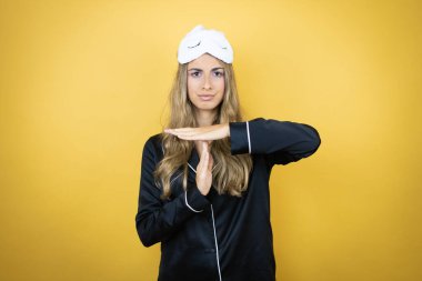 İzole edilmiş sarı arka plan üzerinde uyku maskesi ve pijama giyen genç ve güzel bir kadın.
