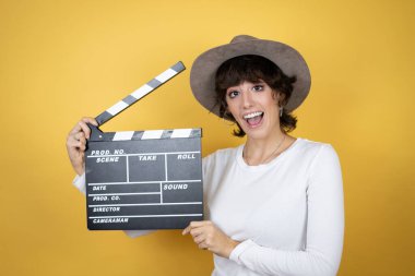 İzole edilmiş sarı arka planda şapka takan beyaz bir kadın elinde klaket tutarak çok mutlu oluyor.